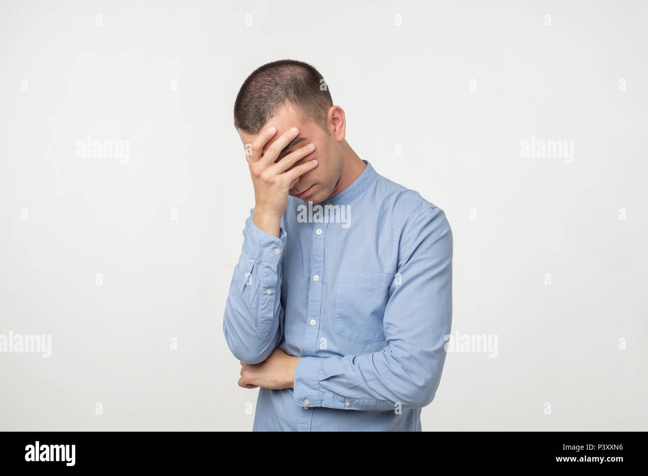 Jeune homme mécontent en chemise bleue couvrant ses yeux sur fond gris. Concept d'émotion négative. Des problèmes au travail ou en famille Banque D'Images Jeune homme mécontent en chemise bleue couvrant ses yeux sur fond gris. Concept d'émotion négative. Des problèmes au travail ou en famille Banque D'Images