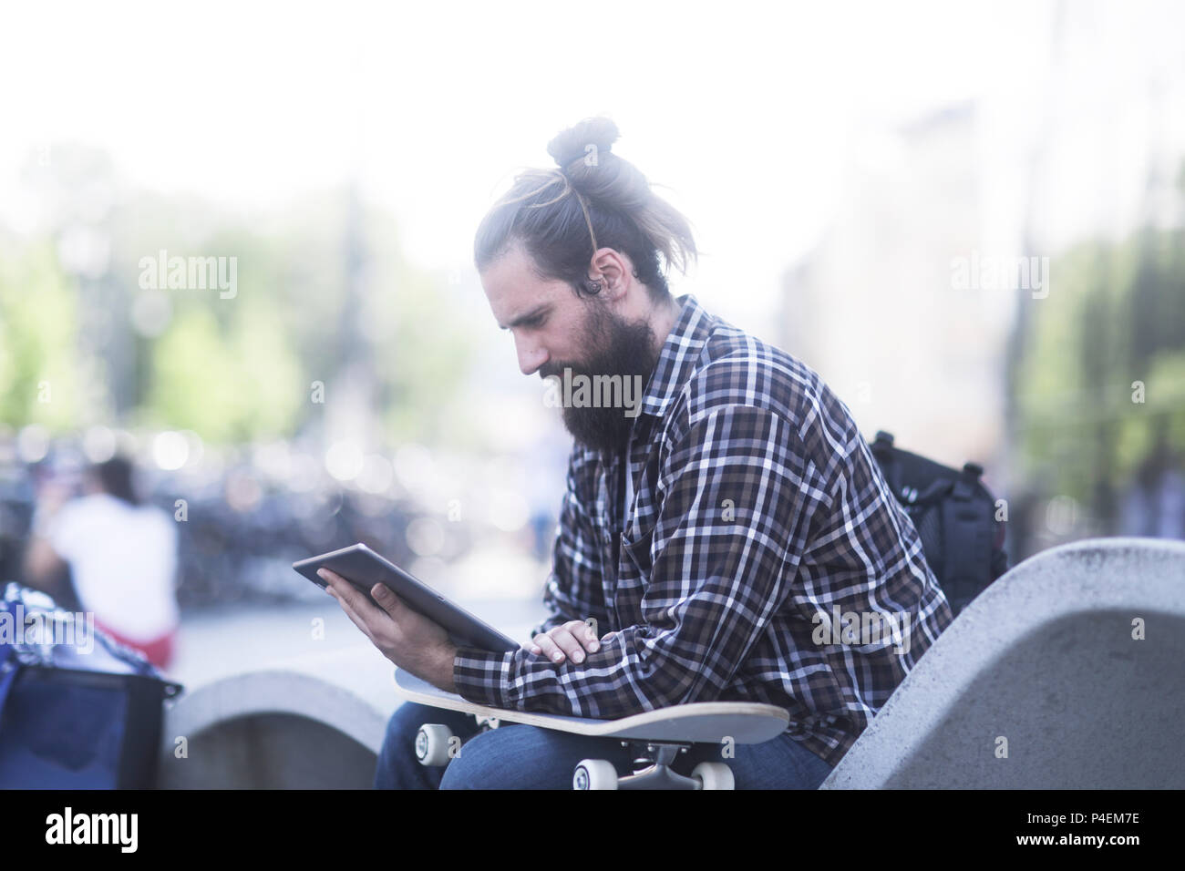 Homme assis sur un banc, s'appuyant sur son skateboard à l'aide d'une tablette numérique Banque D'Images
