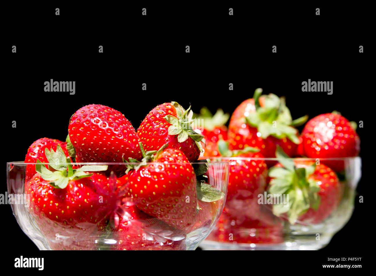 Gros plan de deux plats de dessert en verre de cristal remplis de fraises fraîches juteuses avec feuilles et tiges, fond noir. Assiette de fruits isolée à l'intérieur. Banque D'Images