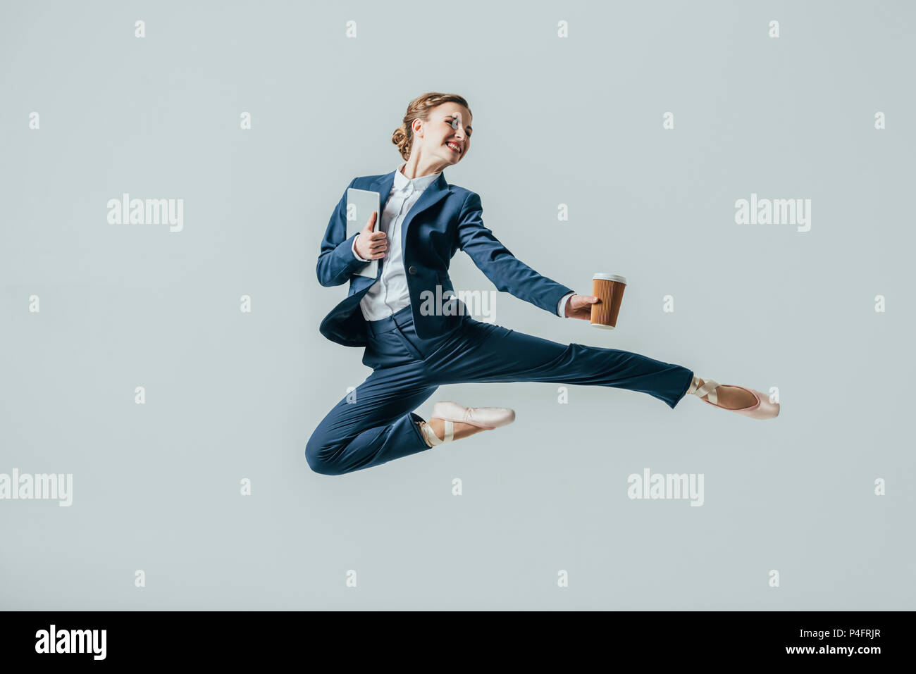 Businesswoman in suit et chaussures de ballet sautant avec café et tablette numérique, isolé sur gris Banque D'Images