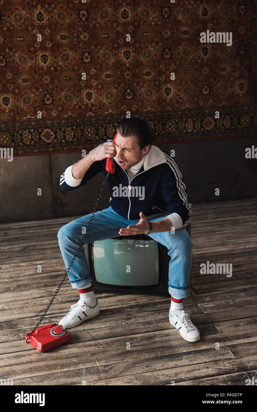 Angry Young Man in vintage clothes sitting on retro tv et parler par téléphone en face de tapis accroché au mur Banque D'Images