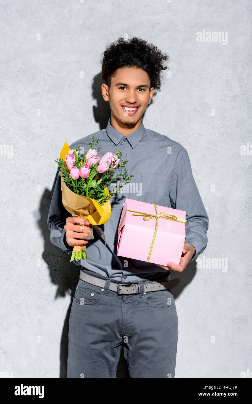 Young African American man holding bouquet de fleurs et de cadeaux sur fond gris Banque D'Images