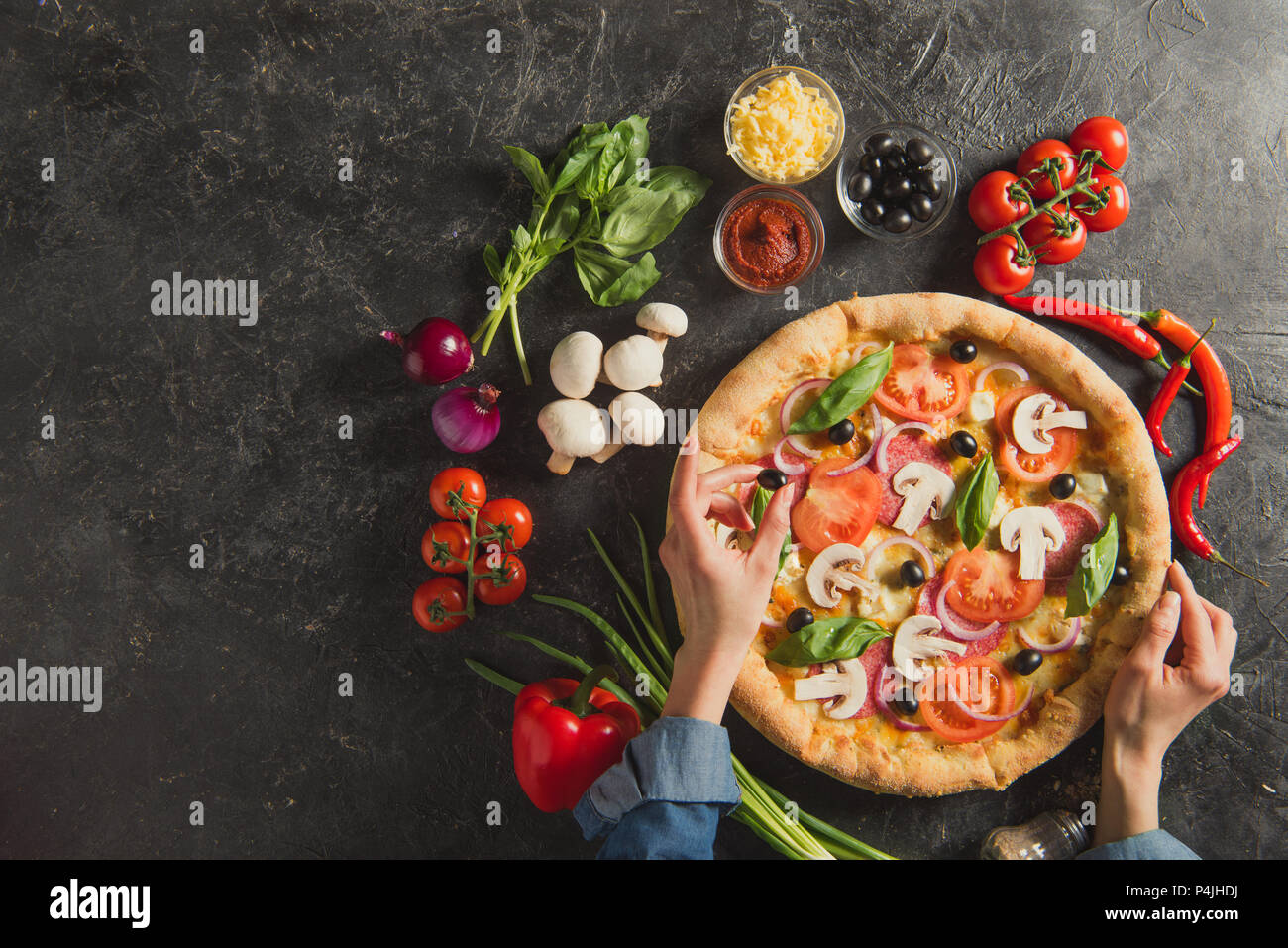 Cropped shot of woman putting olives noires sur la pizza italienne sur la table sombre Banque D'Images