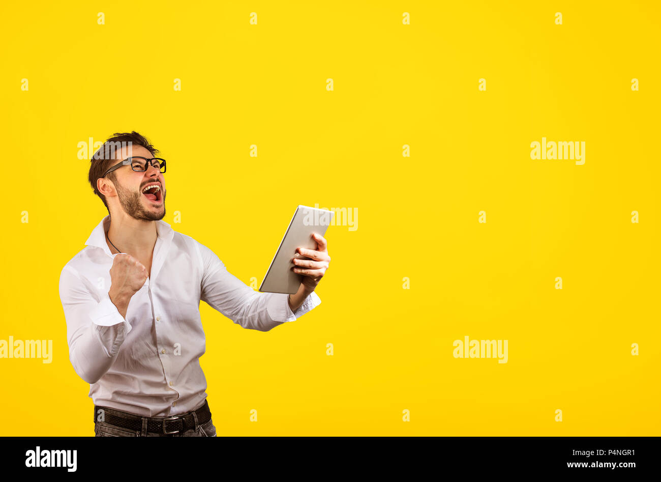Hipster lumineux joyeux man holding fist jusqu'alors que des cris de joie de gagner sur fond jaune et holding tablet Banque D'Images