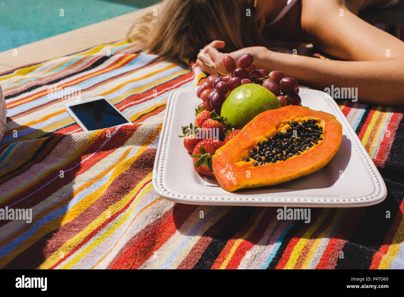 Culture d'une femme sunbaking de la piscine avec un plateau de fruits frais et un iPhone. Banque D'Images