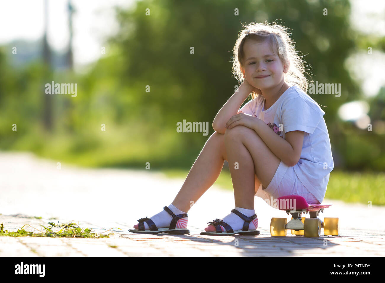 Jolie petite fille blonde en short blanc et T shirt rose assis sur skateboard et souriant en voyant l appareil photo sur un fond d ete brouillee. Enfant Photo Stock Alamy