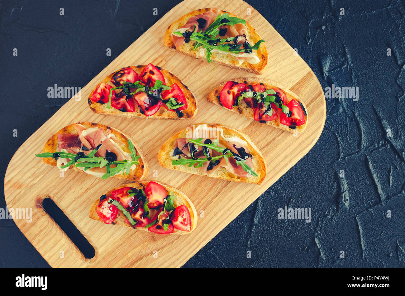 Bruschetta italienne avec tomates, jambon de Parme, roquette et sauce balsamique glasse sur une planche à découper sur pierre sombre arrière-plan avec place pour le texte Banque D'Images