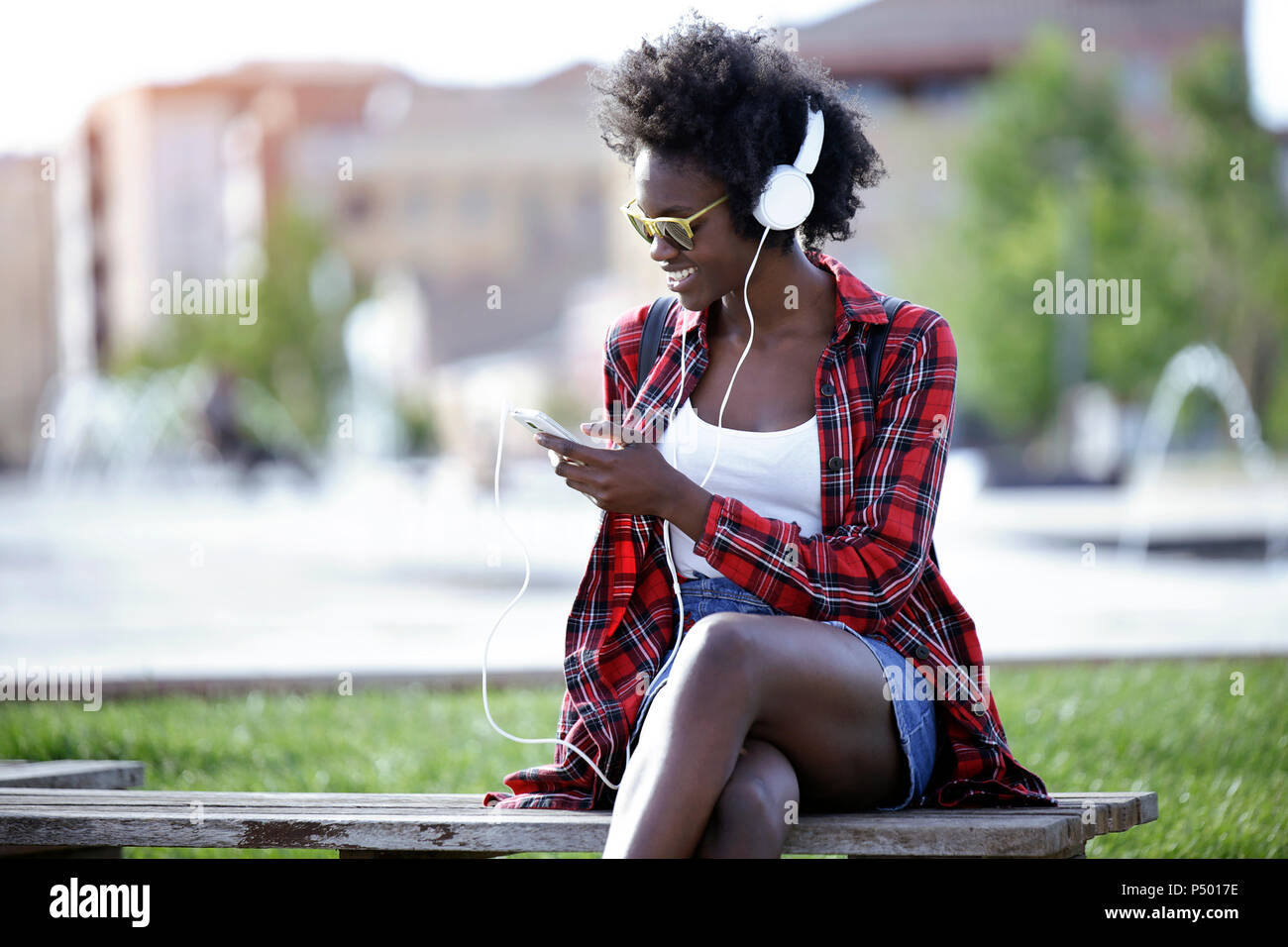 Souriante jeune femme assis sur un banc dans le parc de la ville à l'écoute de la musique avec des écouteurs Banque D'Images