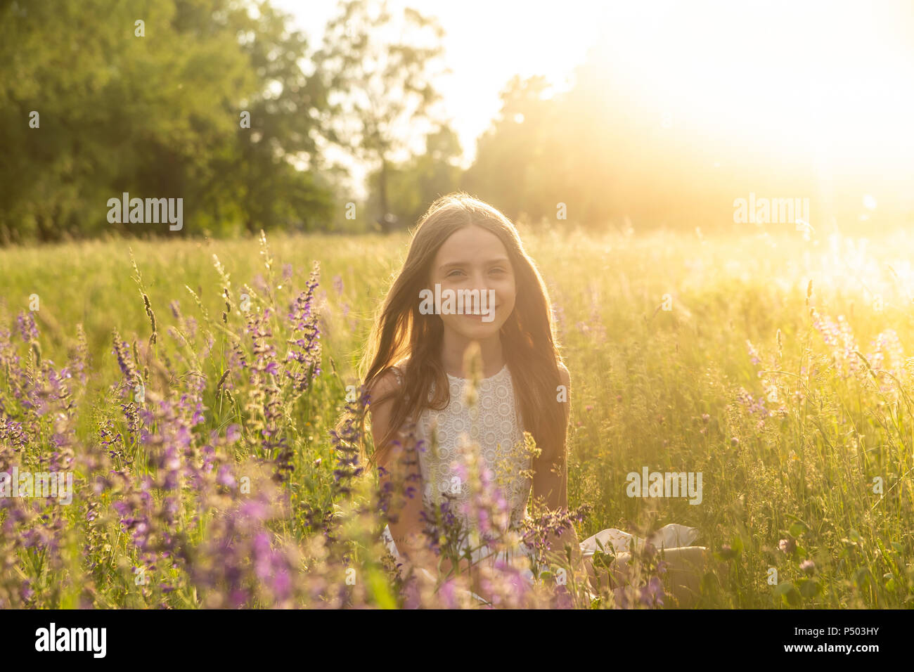 Portrait of smiling girl accroupi sur pré des fleurs au crépuscule du soir Banque D'Images