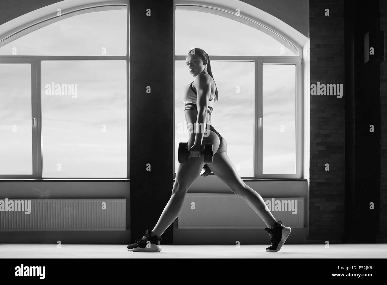 Photo en noir et blanc de la femme sportive avec les décisions des exercices en salle spacieuse avec fenêtres panoramiques. regarde monter, superbes. des muscles puissants. Garder les grandes haltères. Porter des vêtements élégants. Banque D'Images