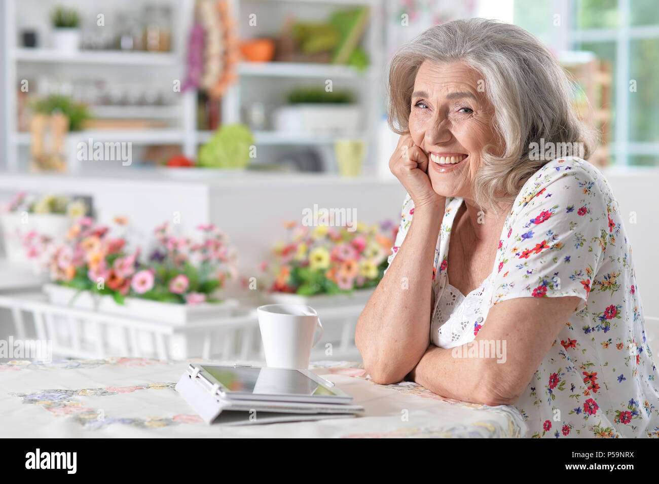 Belle senior woman smiling at camera while drinking tea at table with digital tablet sur elle Banque D'Images
