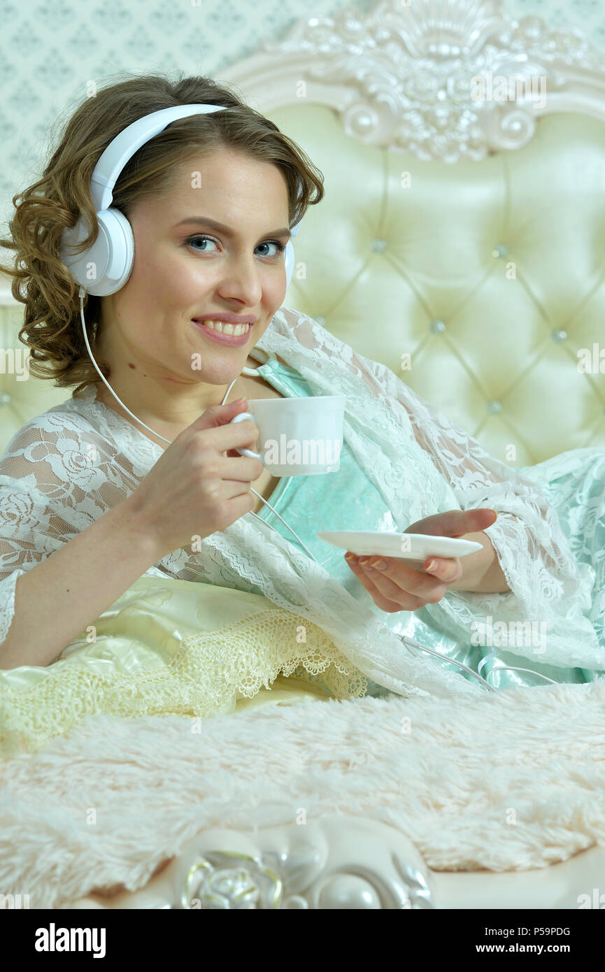 Belle jeune femme à l'écoute de la musique avec des écouteurs et boire le thé au lit Banque D'Images