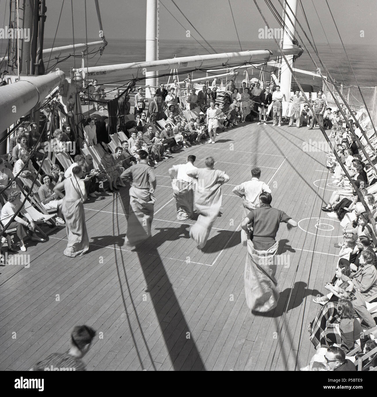 Années 1950, pour passer le temps, s'amuser et rester actif pendant un long voyage, une union-castle steamship voyageant au Cap en Afrique du Sud, les passagers adultes mâles prendre part à un jeu de carte, le sac traditionnel où les concurrents de la course en sacs ou sacs postaux hop depuis un point de départ à une ligne de dimension. D'autres passagers assis dans des transats encourager. Banque D'Images