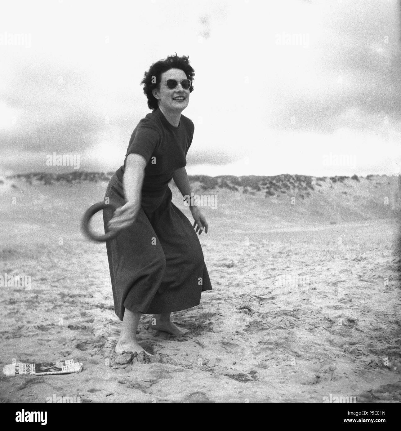Années 1950, belle femme à lunettes à l'extérieur sur une plage de sable à pieds nus, porte une robe de jouer à un jeu de jeter du cercle ou anneau, England, UK. Banque D'Images