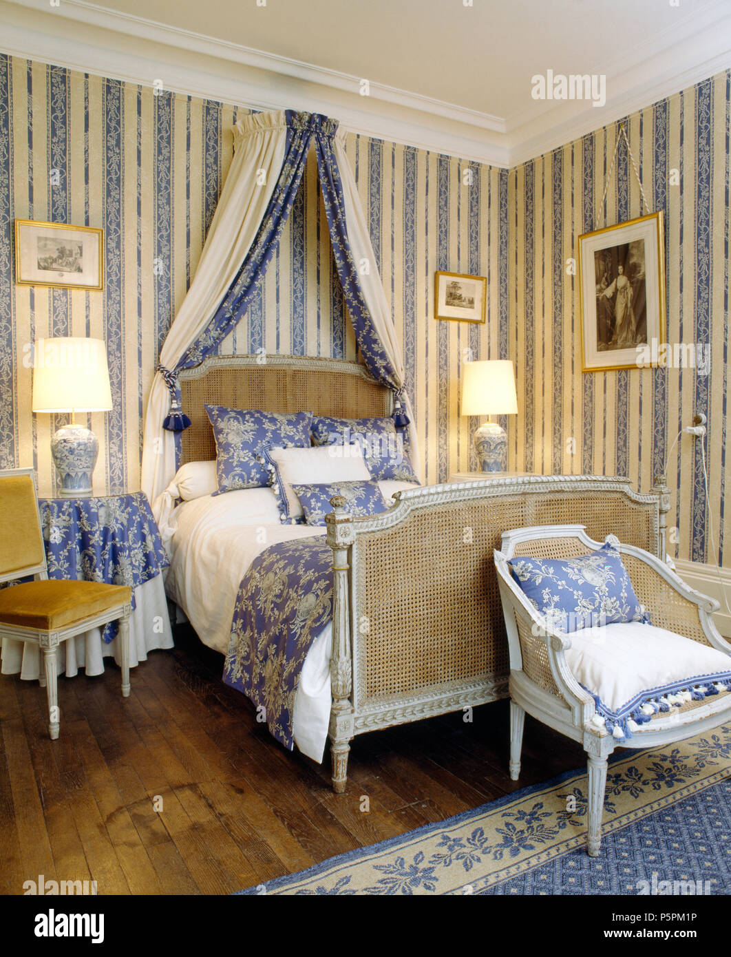 Bergère antique lit avec rideaux bleu en français pays chambre avec un papier peint à rayures blanc +bleu et lampes allumées Banque D'Images