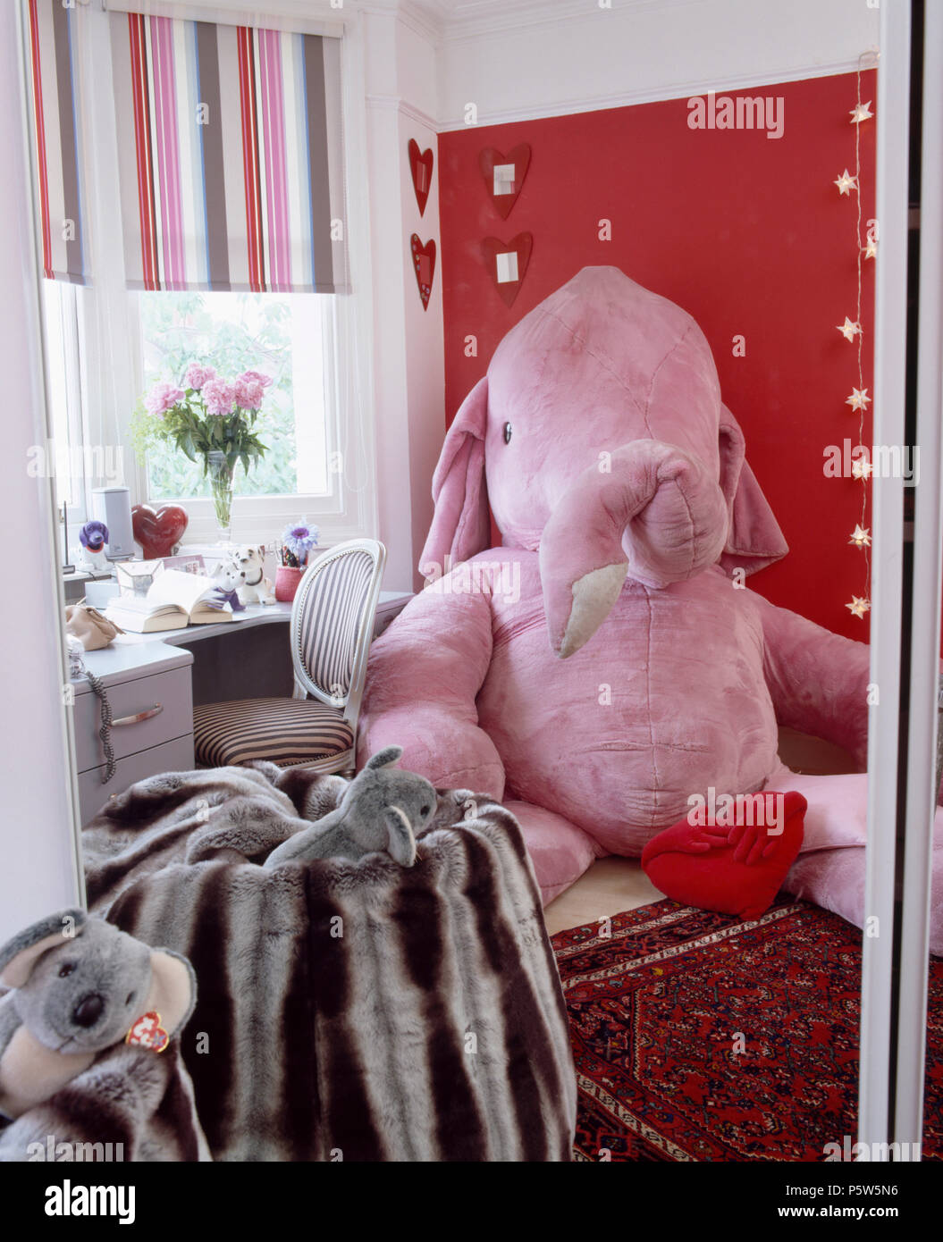 Grand éléphant velours rose contre mur rouge dans la chambre de l'enfant avec un sac de haricots en velours et fenêtre avec rose +rayures gris blind Banque D'Images