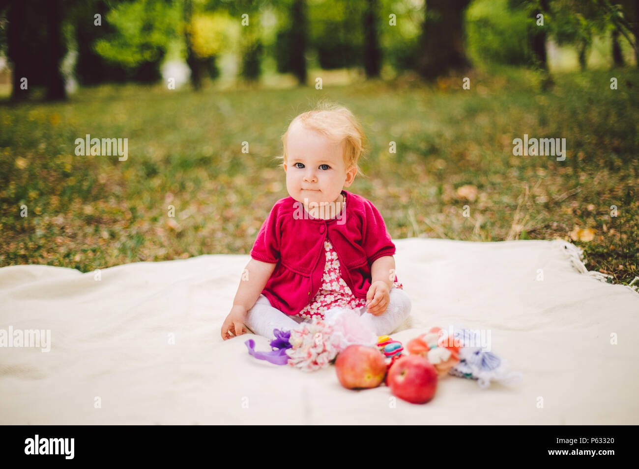 Petite fille blonde Origine ethnique Caucasienne un an à partir de la naissance est assise sur un plaid sur l'herbe verte dans le parc et tient en mains des pommes rouges. Banque D'Images