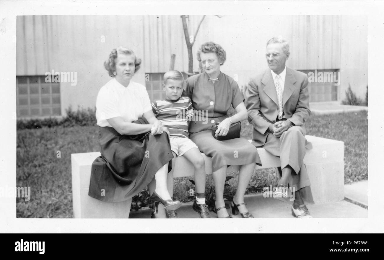 Photographie noir et blanc, montrant une femme avec de courts, frisés, cheveux blonds, portant une jupe et des talons, à l'appareil photo, au sérieux tout en étant assis sur un banc en béton à côté d'un garçon blond portant une chemise rayée et shorts, également vêtu d'un air sérieux, avec un couple plus âgé habillé formellement (vraisemblablement le boy's grands-parents) à droite, et un bâtiment en béton avec windows en blocs de verre visible à l'arrière-plan, probablement photographié dans l'Ohio, 1955. () Banque D'Images