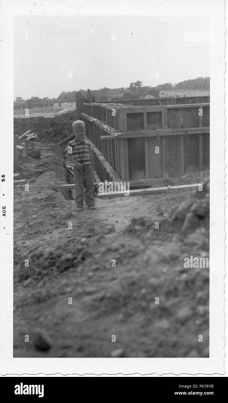 Photographie noir et blanc, montrant un petit garçon aux cheveux blonds, face à l'appareil photo en vue de pleine longueur, vêtu d'une chemise rayée, pantalons et chaussures, debout au milieu d'un site de construction, avec l'ossature en bois, la saleté et les débris de construction visible à l'arrière-plan, probablement photographié dans l'Ohio, Août, 1956. () Banque D'Images