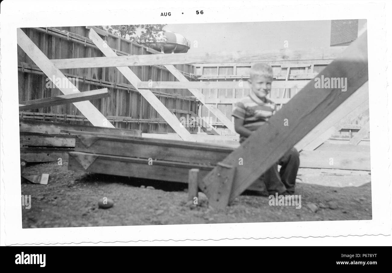 Photographie noir et blanc, montrant un petit garçon aux cheveux blonds, portant une chemise rayée, et souriant à la caméra en position assise sur plusieurs longueurs de bois en appui sur le sol de terre, entourée d'éléments de charpente, avec un château d'eau visible à l'arrière-plan, probablement photographié dans l'Ohio, Août, 1956. () Banque D'Images