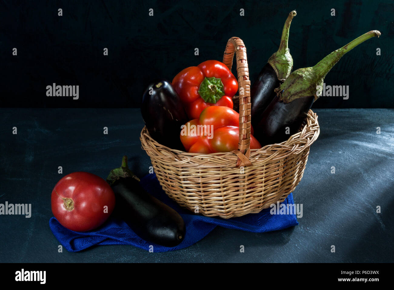 Les légumes en panier d'osier et de l'aubergine et tomate sur serviette bleue sur fond sombre. Banque D'Images