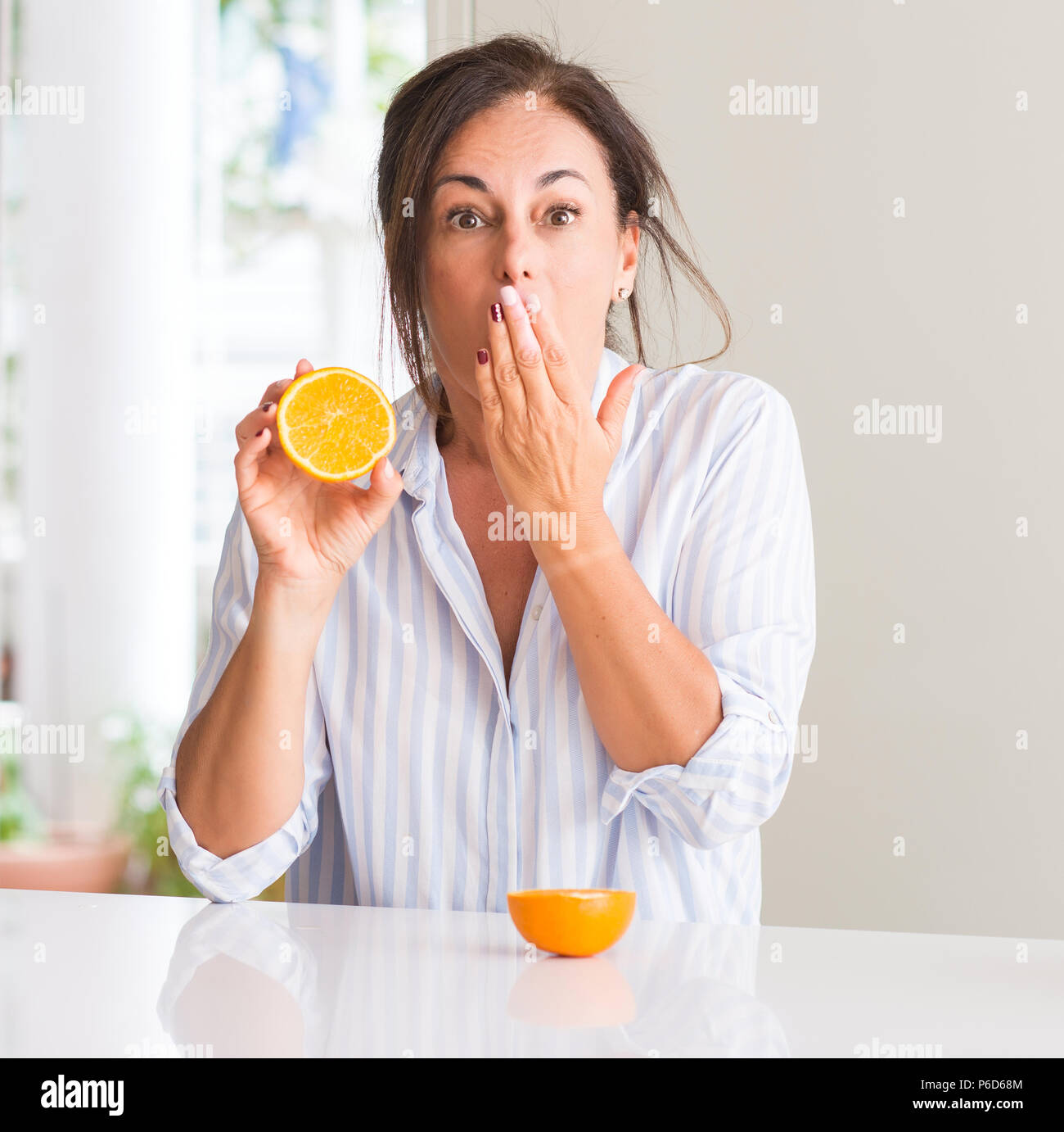 Middle aged woman holding orange fruit couvrir la bouche à part choqué avec honte pour erreur, expression de la peur, peur en silence, concept secret Banque D'Images