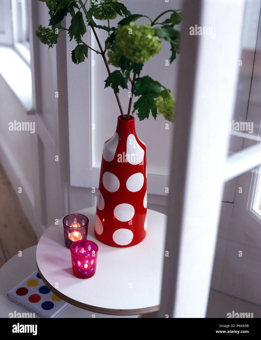 Vase tachetée rouge et blanc avec le feuillage sur un petit tableau blanc avec des bougies allumées en rouge les détenteurs de verre Banque D'Images