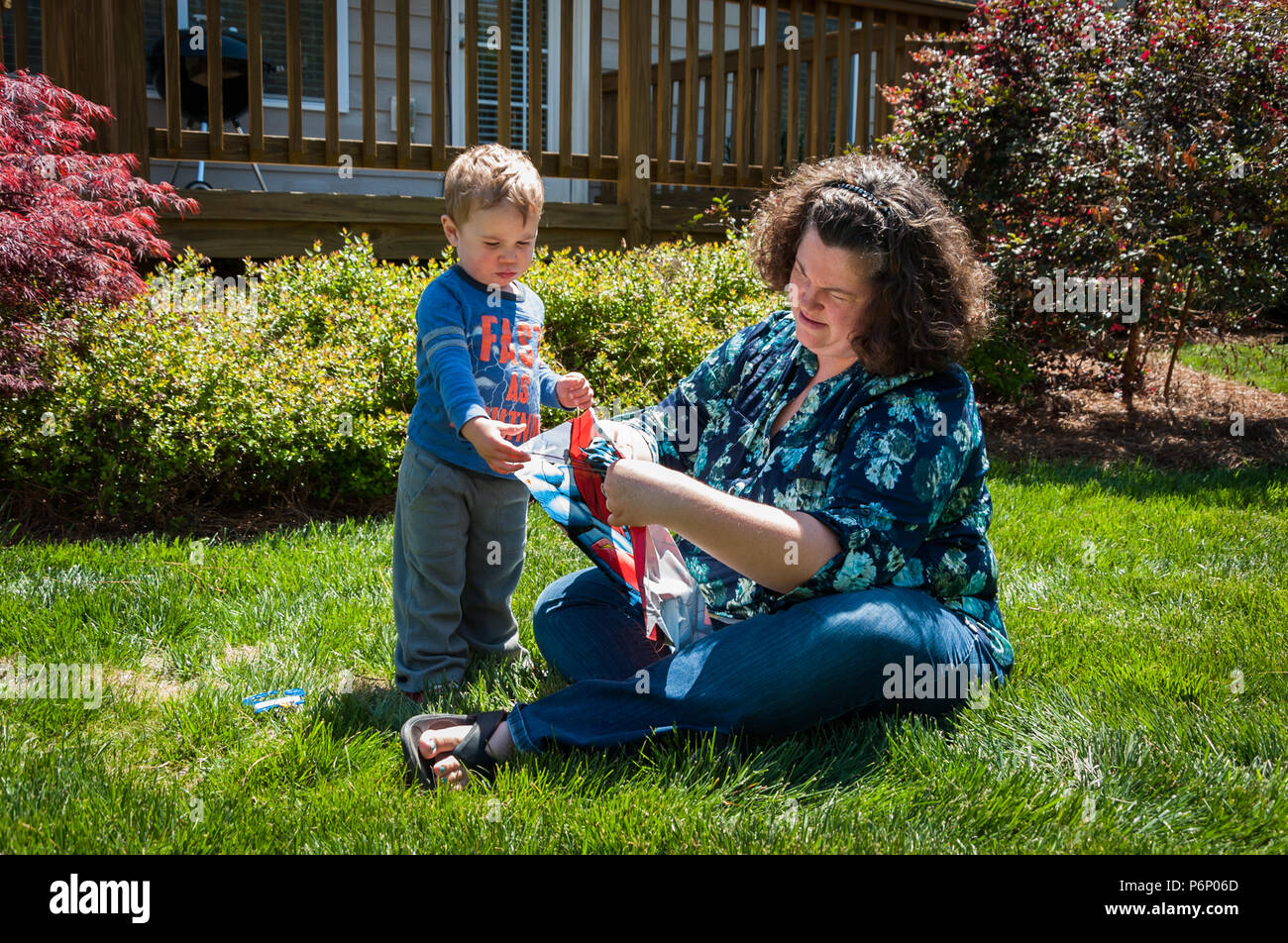 Charlotte, NC-Avril 17, 2016 : Jeune garçon regarde sa mère monter un cerf-volant dans la cour. Mère est assise dans l'herbe alors que le jeune fils tient sa main. Banque D'Images