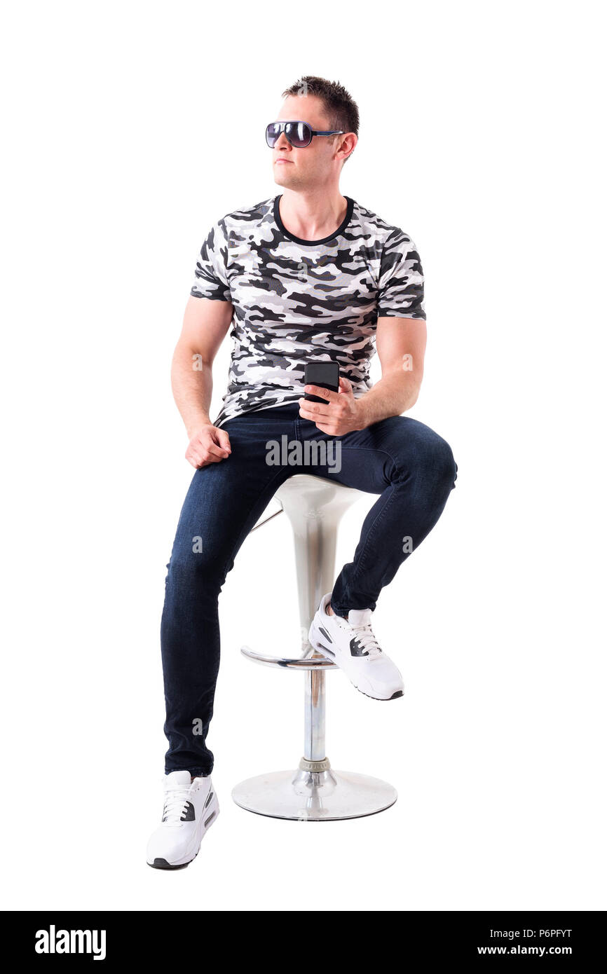 Cool man wearing sunglasses et camouflage t-shirt holding mobile phone à la route assis sur tabouret de bar. Corps plein isolé sur fond blanc. Banque D'Images