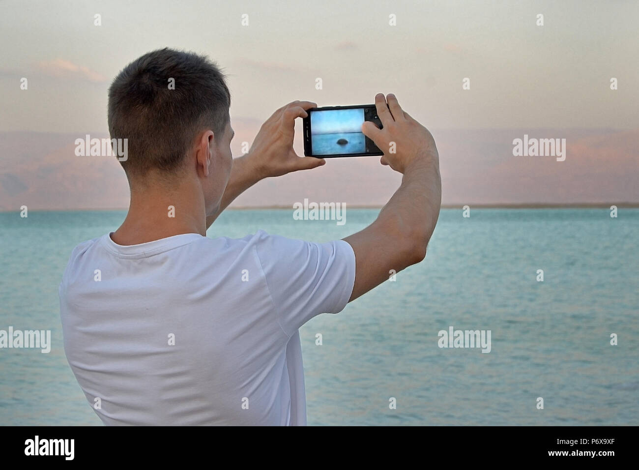 Guy touriste dans un T-shirt blanc prend des photos à l'aide d'une pierre du smartphone dans la mer au coucher du soleil. La mer Morte, en Israël. Paysage de montagne de rêve de Jordanie Banque D'Images