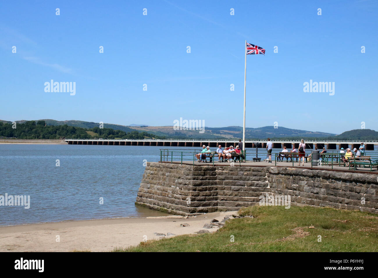 Les gens sur le quai à Arnside par le fleuve de l'estuaire de Kent à marée montante avec Arnside viaduc de chemin de fer derrière le drapeau de l'Union et sur le mât. Banque D'Images