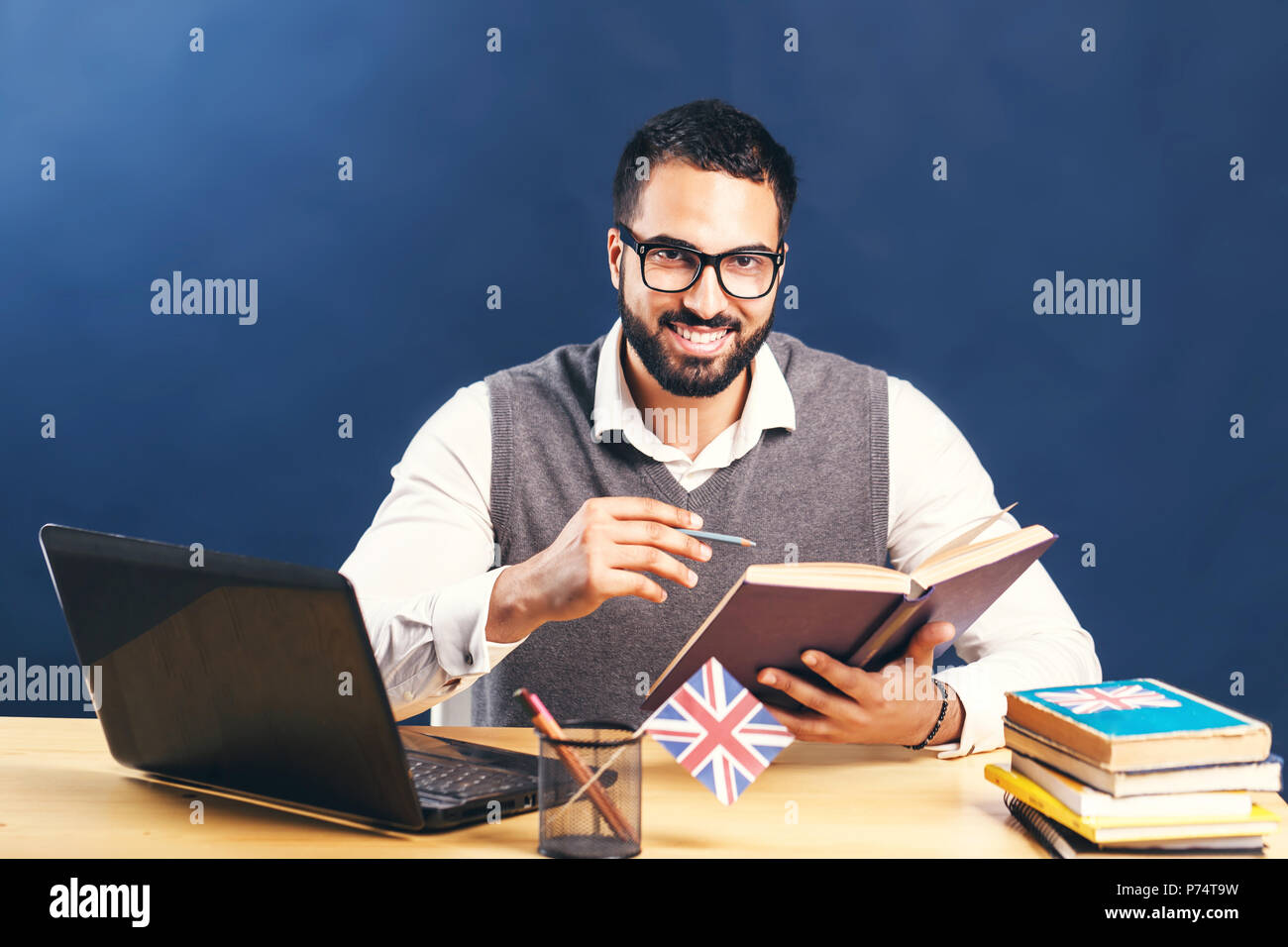Homme barbu aux cheveux noirs l'apprentissage de l'anglais, le port de chandail gris veste et chemise blanche immaculée, travailler dur à l'office de bureau avec ordinateur portable avant mur noir Banque D'Images