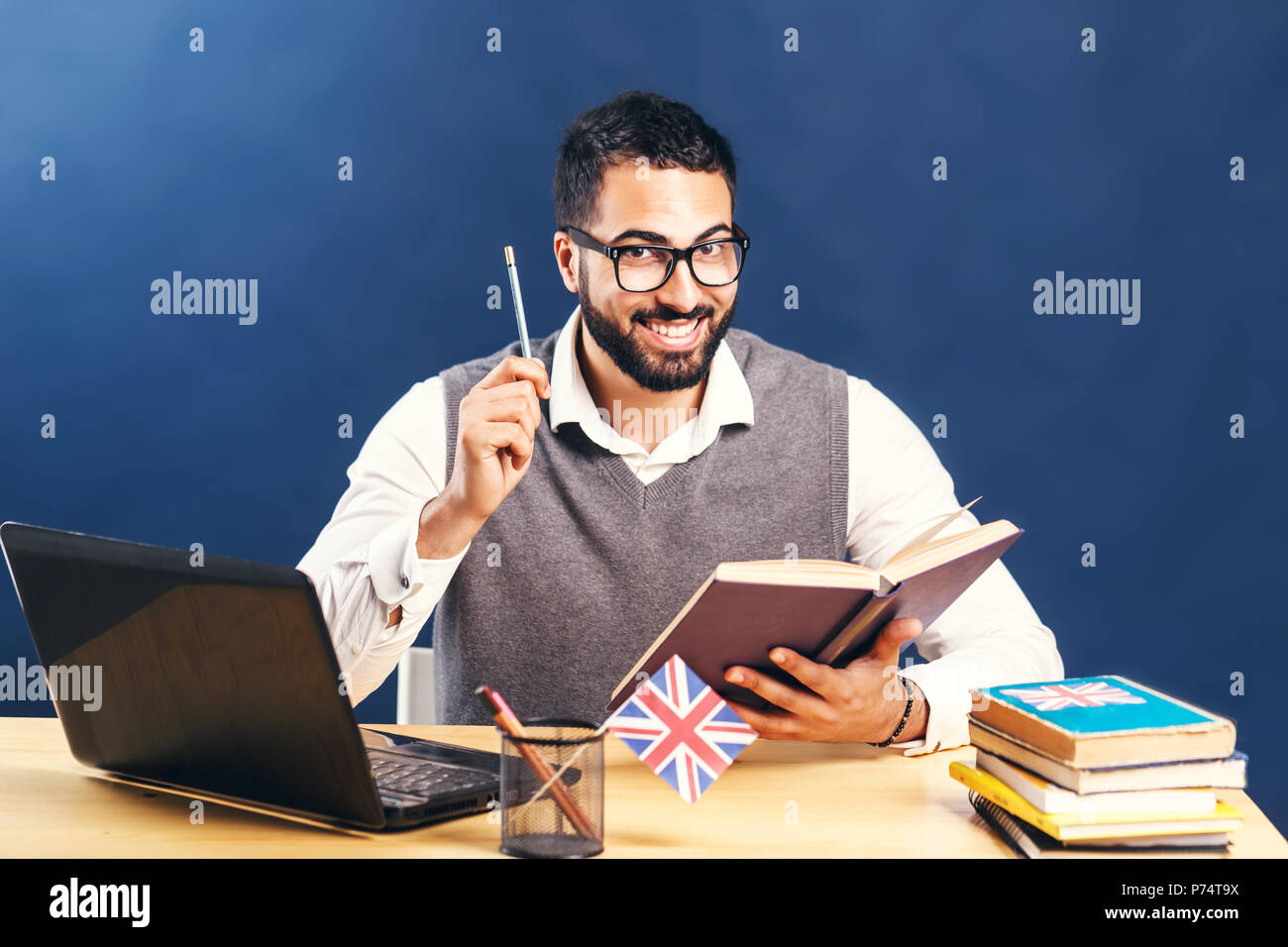 L'homme aux cheveux noirs l'apprentissage de l'anglais, le port de chandail gris veste et chemise blanche immaculée, souriant à l'office de bureau avec ordinateur portable avant mur noir Banque D'Images
