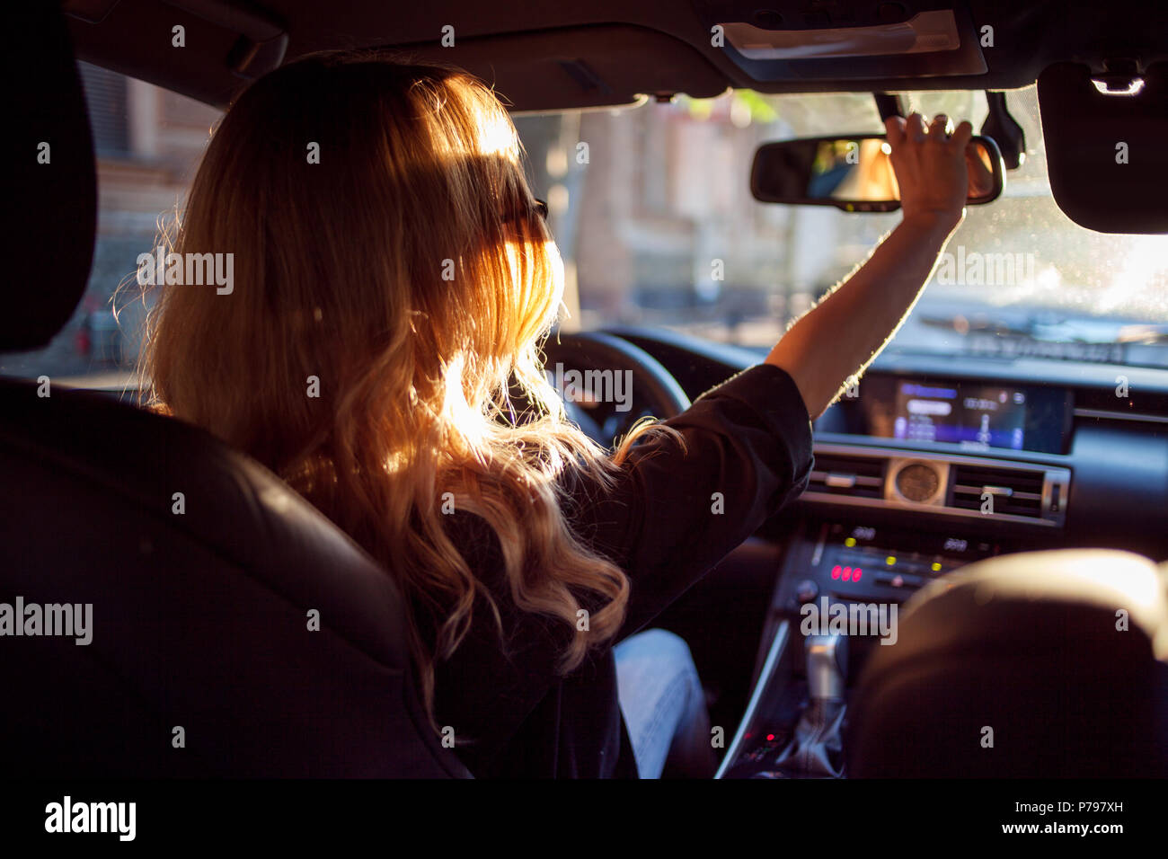 Belle jeune femme à lunettes assis derrière le volant d'une voiture et  corrige miroir. Photo à partir de la banquette arrière. Plein soleil dans le  f Photo Stock - Alamy