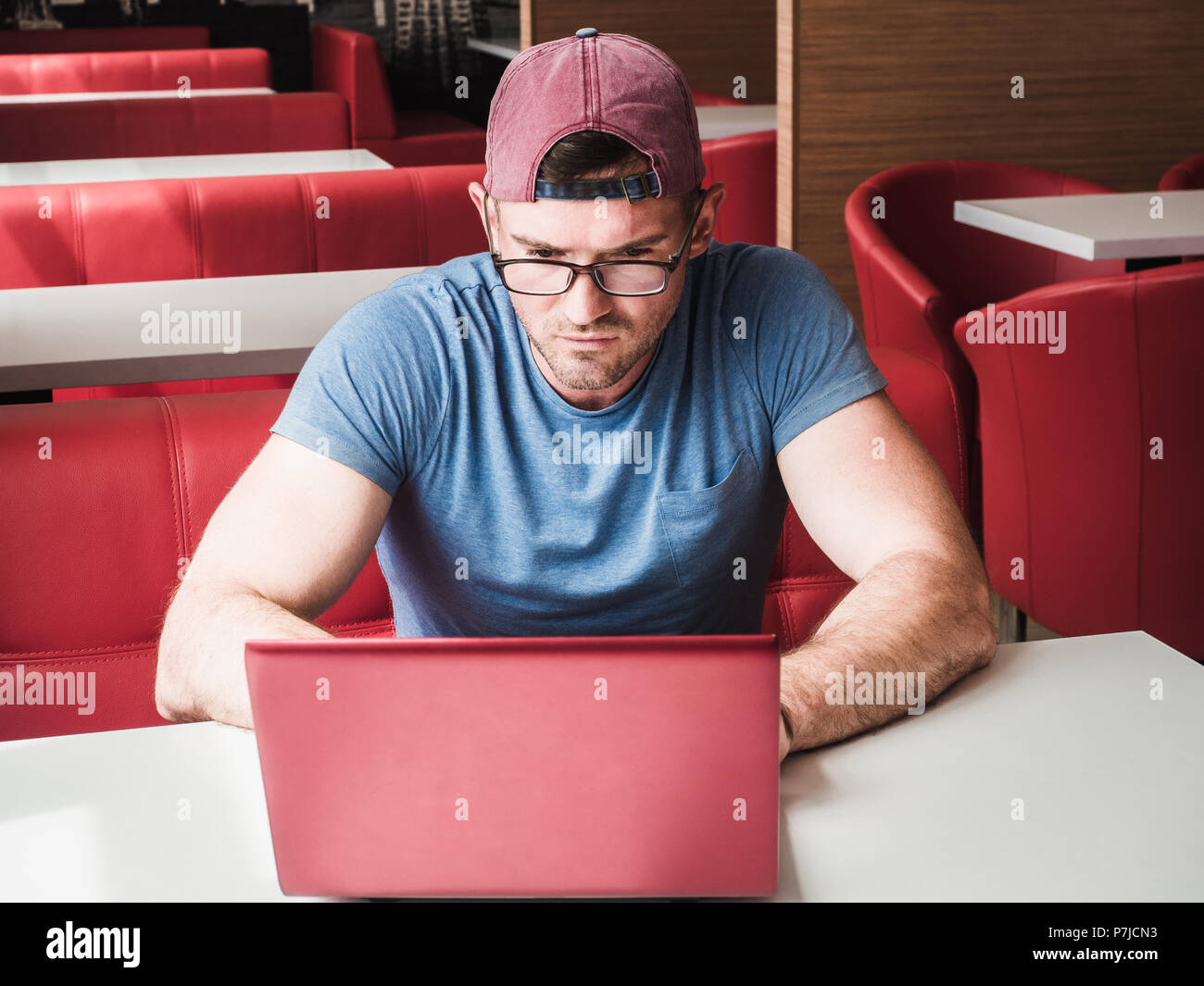 Beau, élégant, sérieux mec avec lunettes, casquette et tee-shirt bleu,  assise seule dans un bar vide sur une chaise rouge et de travail sur  l'ordinateur à l'étude whi Photo Stock - Alamy