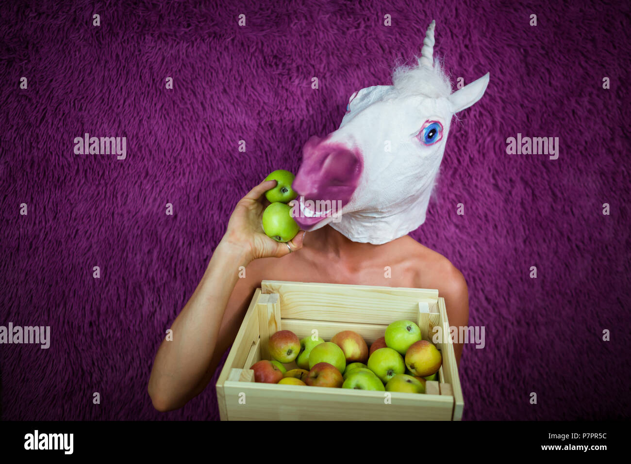 Freaky jeune femme au masque comique se dresse sur fond violet avec la boîte de fruits. Portrait de Dame inhabituelle. Funny Girl licorne détient des pommes dans la main. Banque D'Images