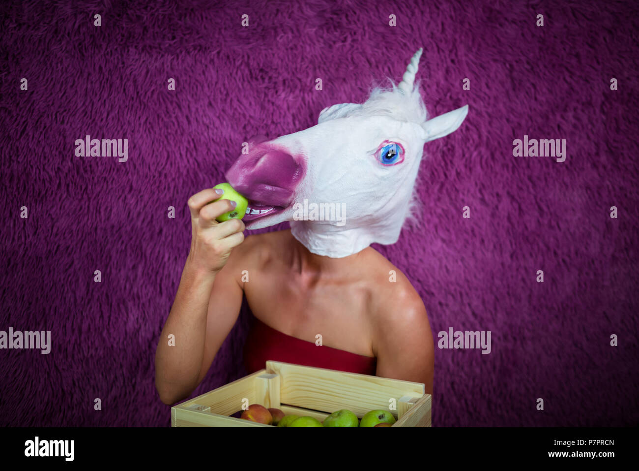 Freaky jeune femme au masque comique se tient sur le fond violet et montre fort de fruits. Funny Girl licorne manger apple. Portrait de Dame inhabituelle. Banque D'Images