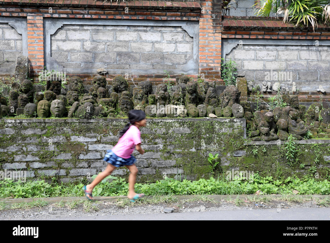 Ubud, Indonésie - le 28 juin 2018 : Une jeune fille court le long d'une voie passé une collection de masques en pierre, à la périphérie de Ubud sur l'île de Bali, Indon Banque D'Images
