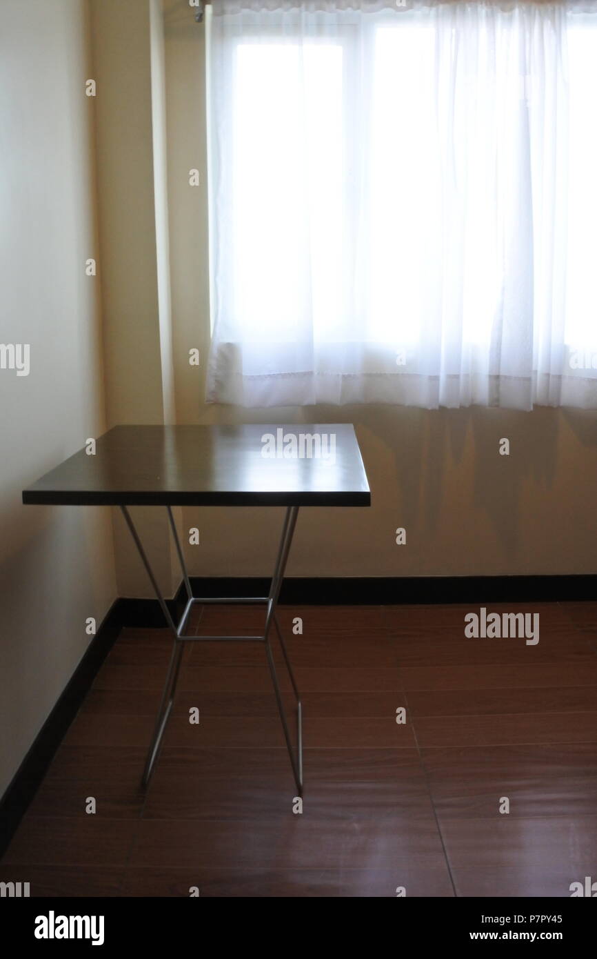 Une salle à l'éclairage tamisé avec lumière naturelle diffusant à travers une fenêtre à rideaux blancs, présentant une table d'angle contre un plancher en bois. Banque D'Images