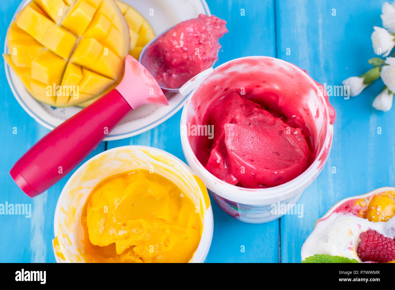 La crème glacée aux fruits à la mangue et les framboises dans un seau. Cuillère pour la crème glacée. Fond bleu et fleurs. copier l'espace. Banque D'Images