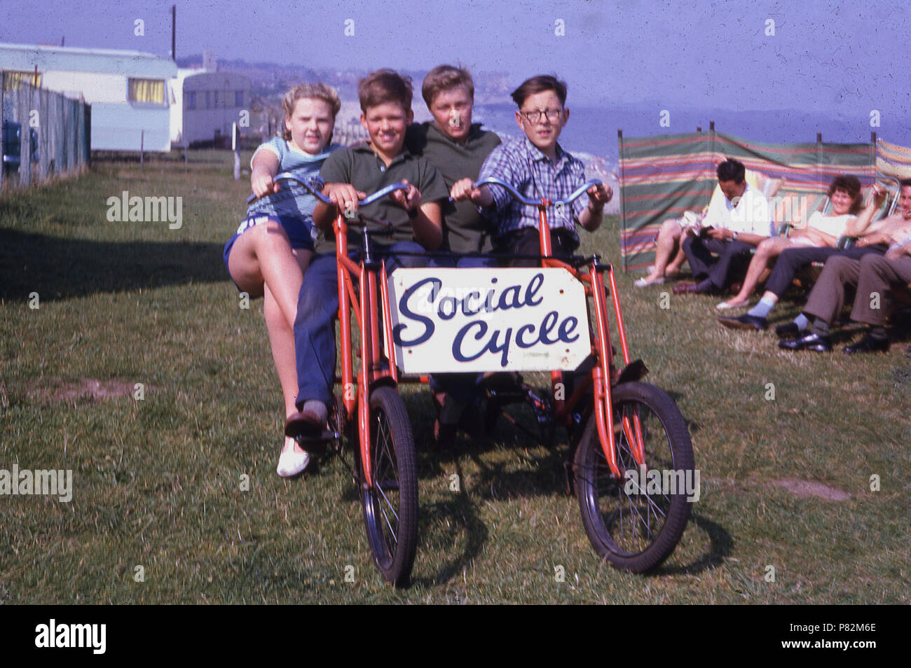 Années 1950, quatre jeunes à l'extérieur assis sur une personne deux 'cycle', à une maison de vacances côtières ou Caravan Park, Angleterre, Royaume-Uni, avec leurs parents au repos dans les transats. Banque D'Images