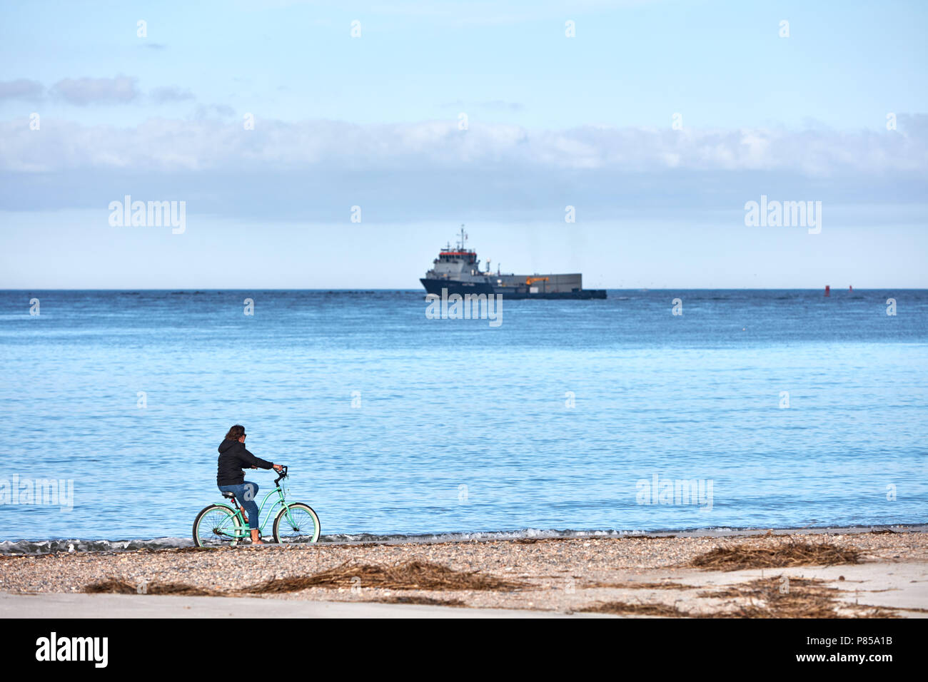 Femme de faire du vélo sur la plage comme un navire de la Marine américaine passé Banque D'Images