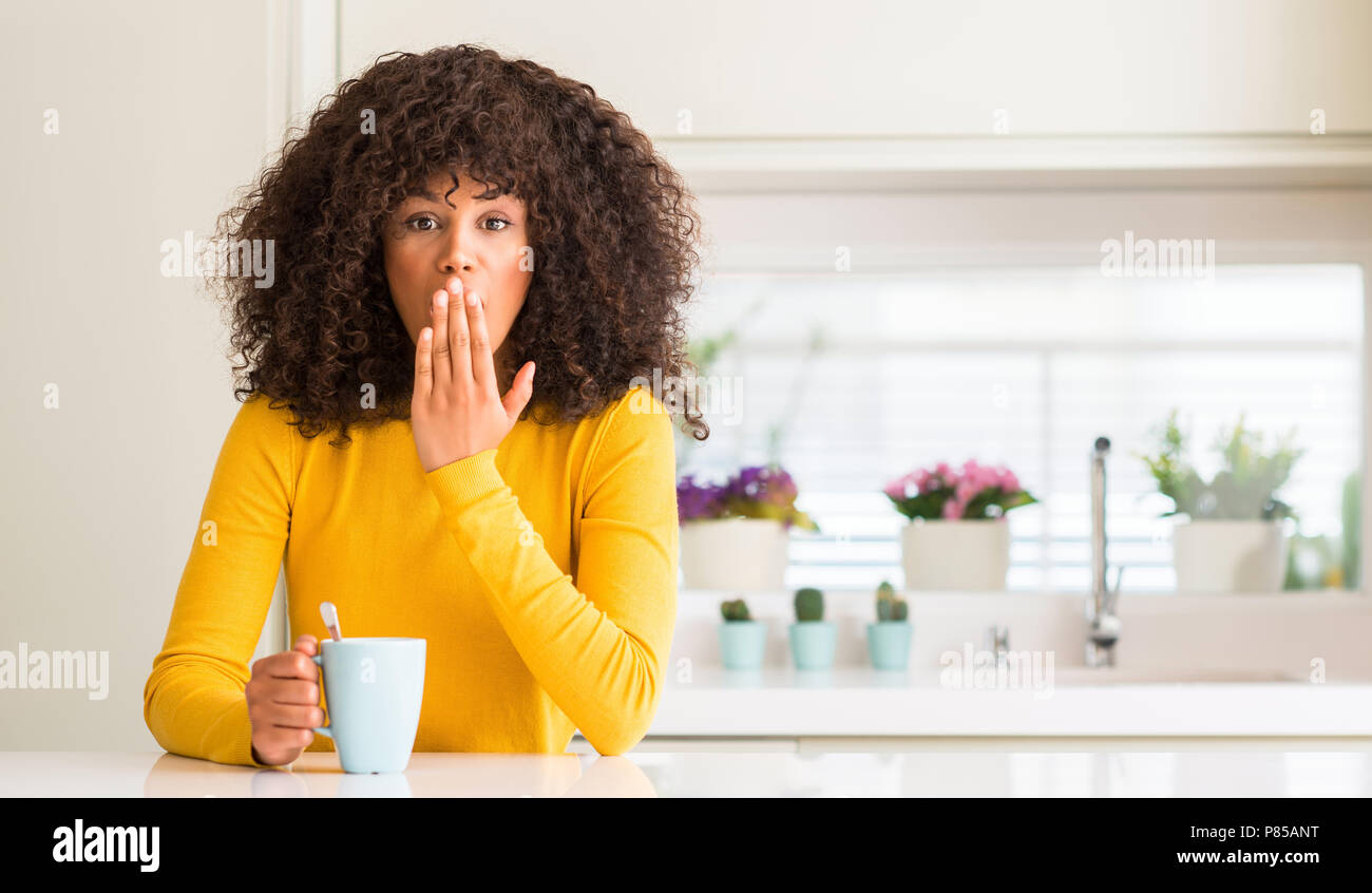 Belle african american woman holding une tasse de café à la maison couvrir la bouche à part choqué avec honte pour erreur, expression de la peur, peur de l'al. Banque D'Images