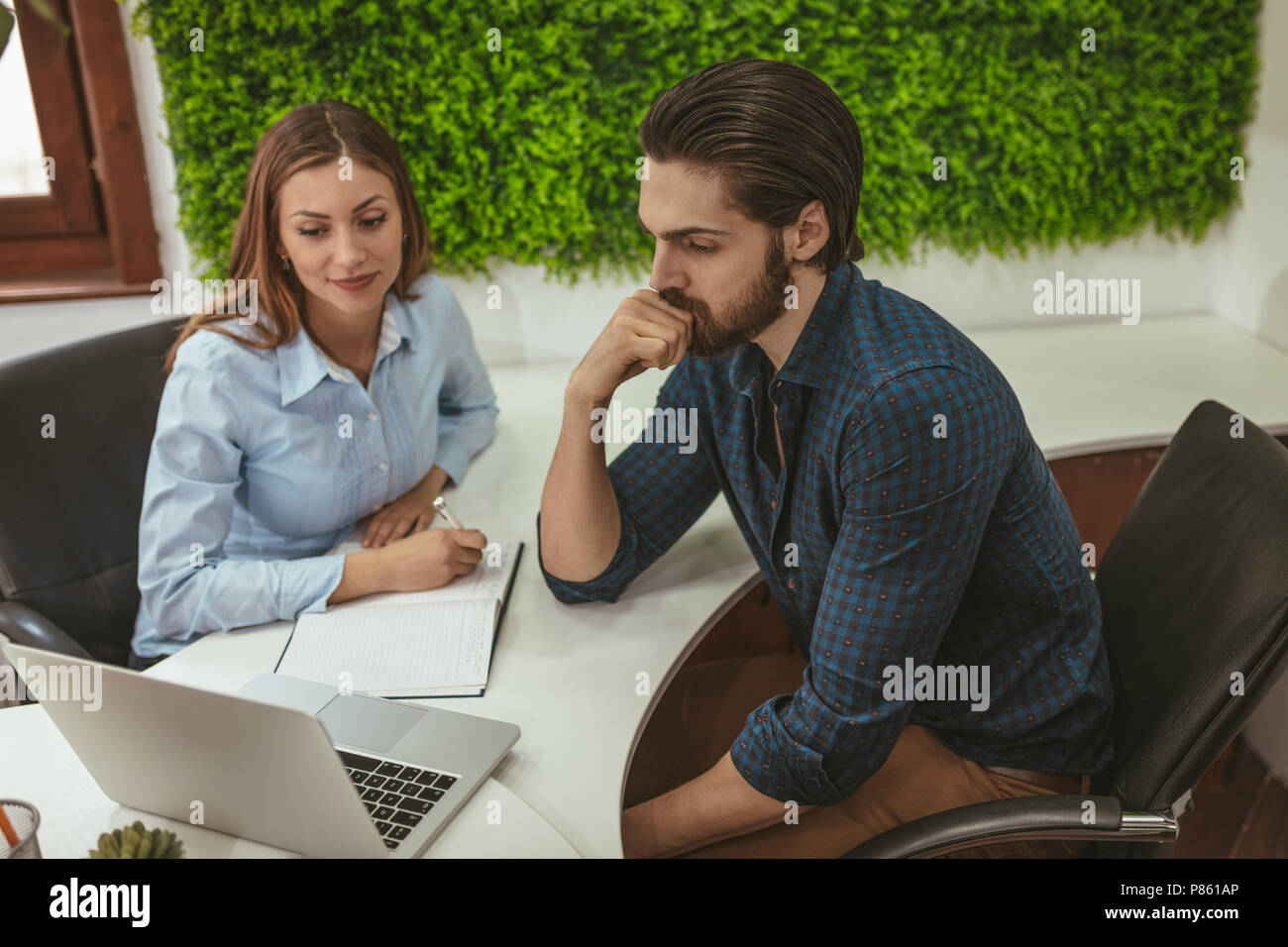 Young handsome stern pensive businessman working on laptop au bureau et jeune femme écoute son collègue et à la recherche à l'écran. Banque D'Images