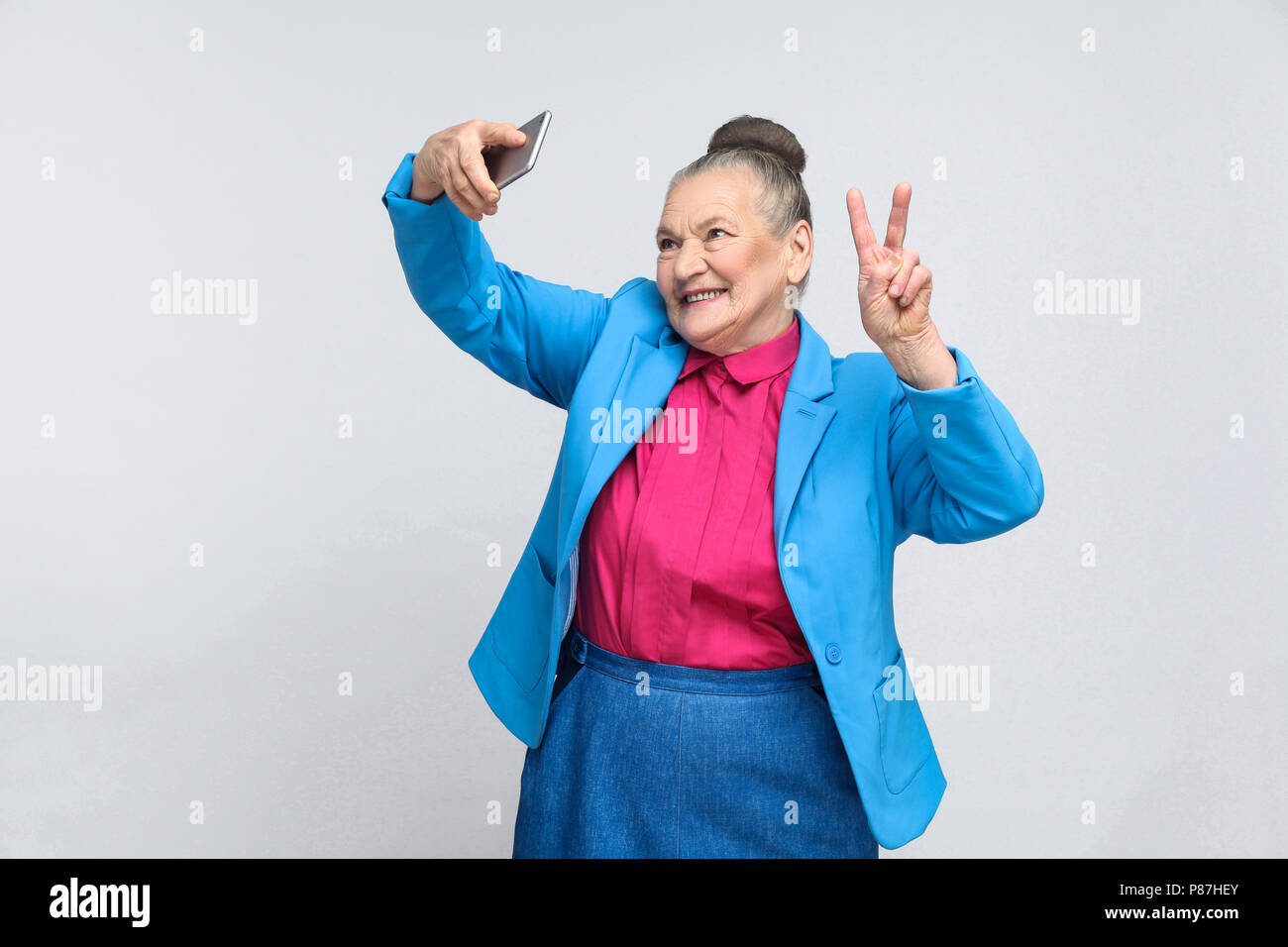 De femme faisant chanter la paix avec selfies et souriant. Beau Portrait de grand-mère expressive en bleu clair costume avec recueillies cheveux gris bun hai Banque D'Images