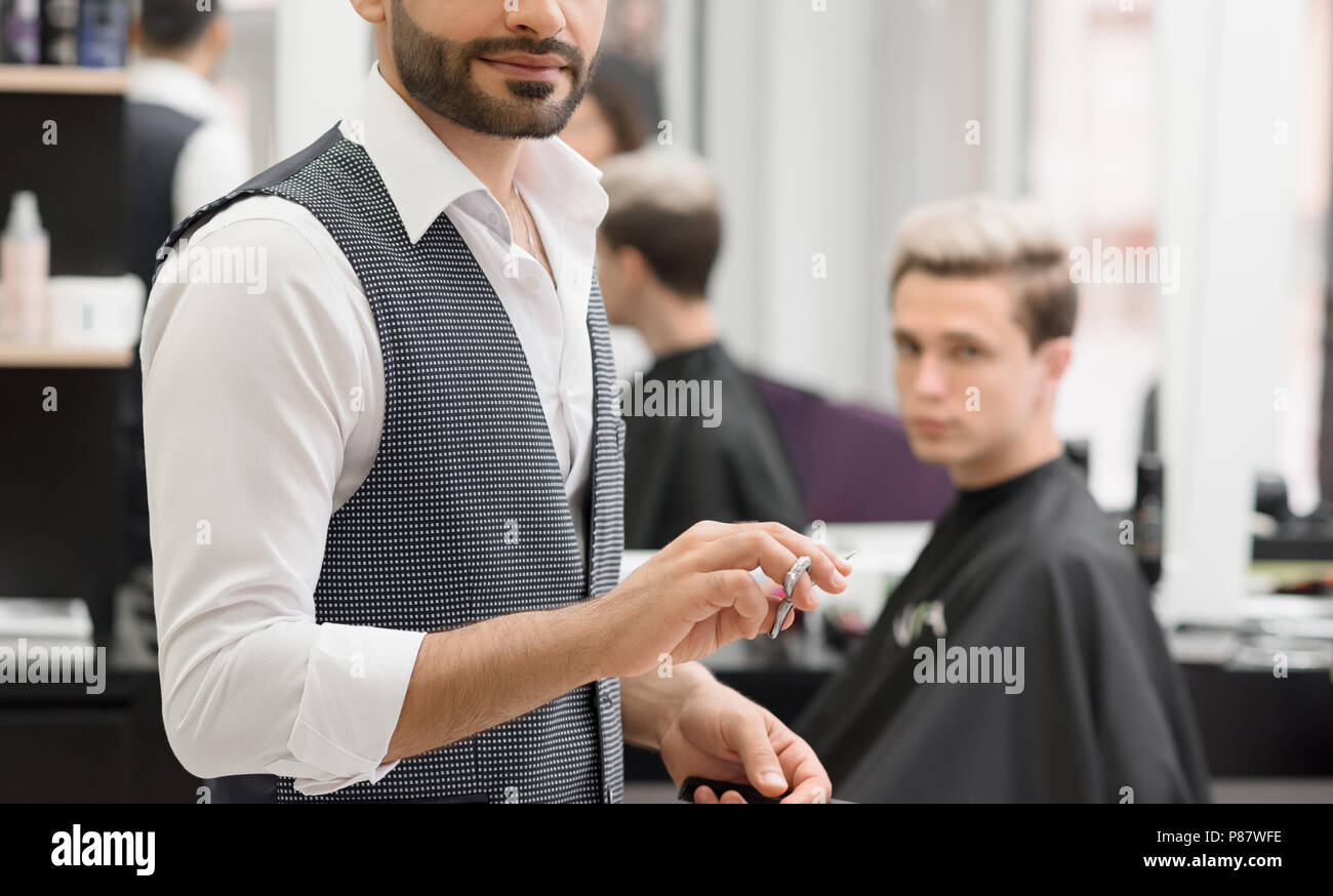 Photo recadrée de coiffure travaillant dans l'aide de ciseaux de coiffure. Vêtu de blanc classic shirt, gilet gris. Décisions à la mode, coupe élégante fo Banque D'Images