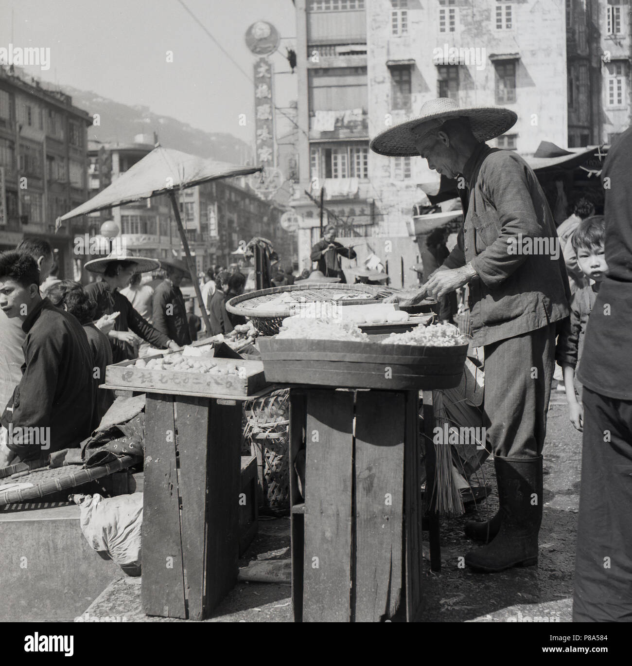 Années 1950, central hong kong, négociant de la rue de sexe masculin, le port d'un chapeau conique ou coolie de marcher sur le soleil chaud, prépare son food pour la journée à venir. Banque D'Images