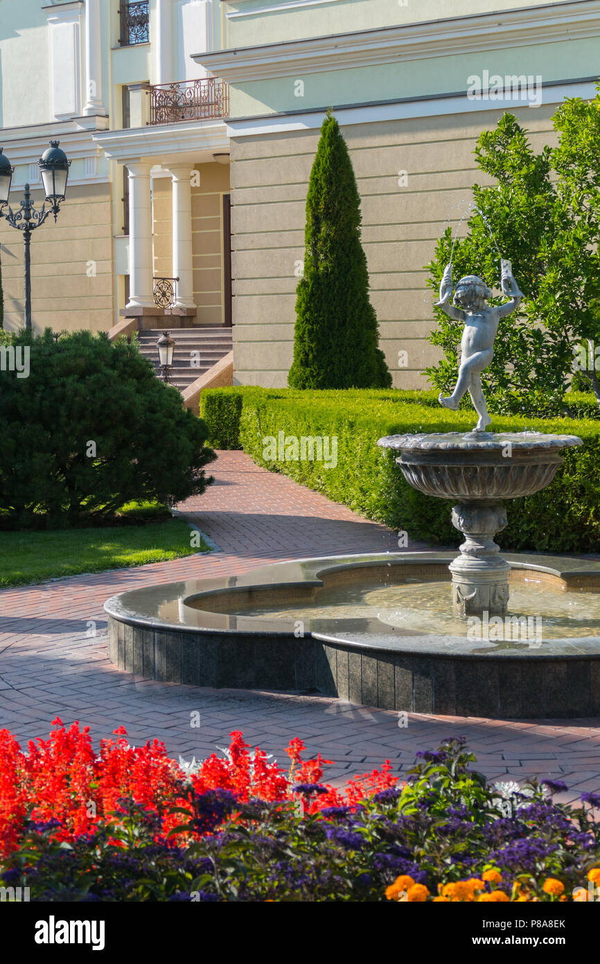 Fontaine avec une statue d'un enfant danse sur un fond d'arbustes parfaitement coupés et de belles fleurs rouges vives . Pour votre conception Banque D'Images