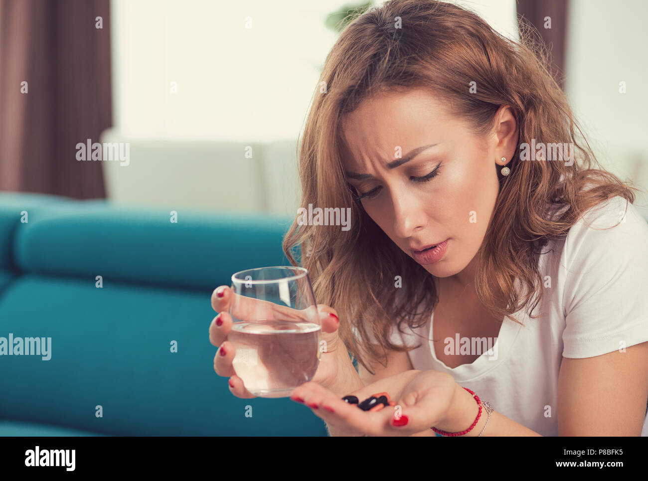 Souligné déprimé attractive young woman avec pilules antidouleur et verre de l'eau assis sur un canapé dans son appartement Banque D'Images