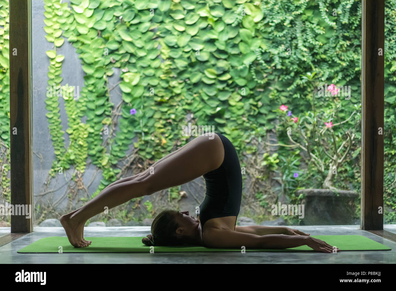 Vue latérale du jeune sportive woman practicing yoga in charrue Halasana (posture) en face de mur recouvert avec des feuilles vertes Banque D'Images